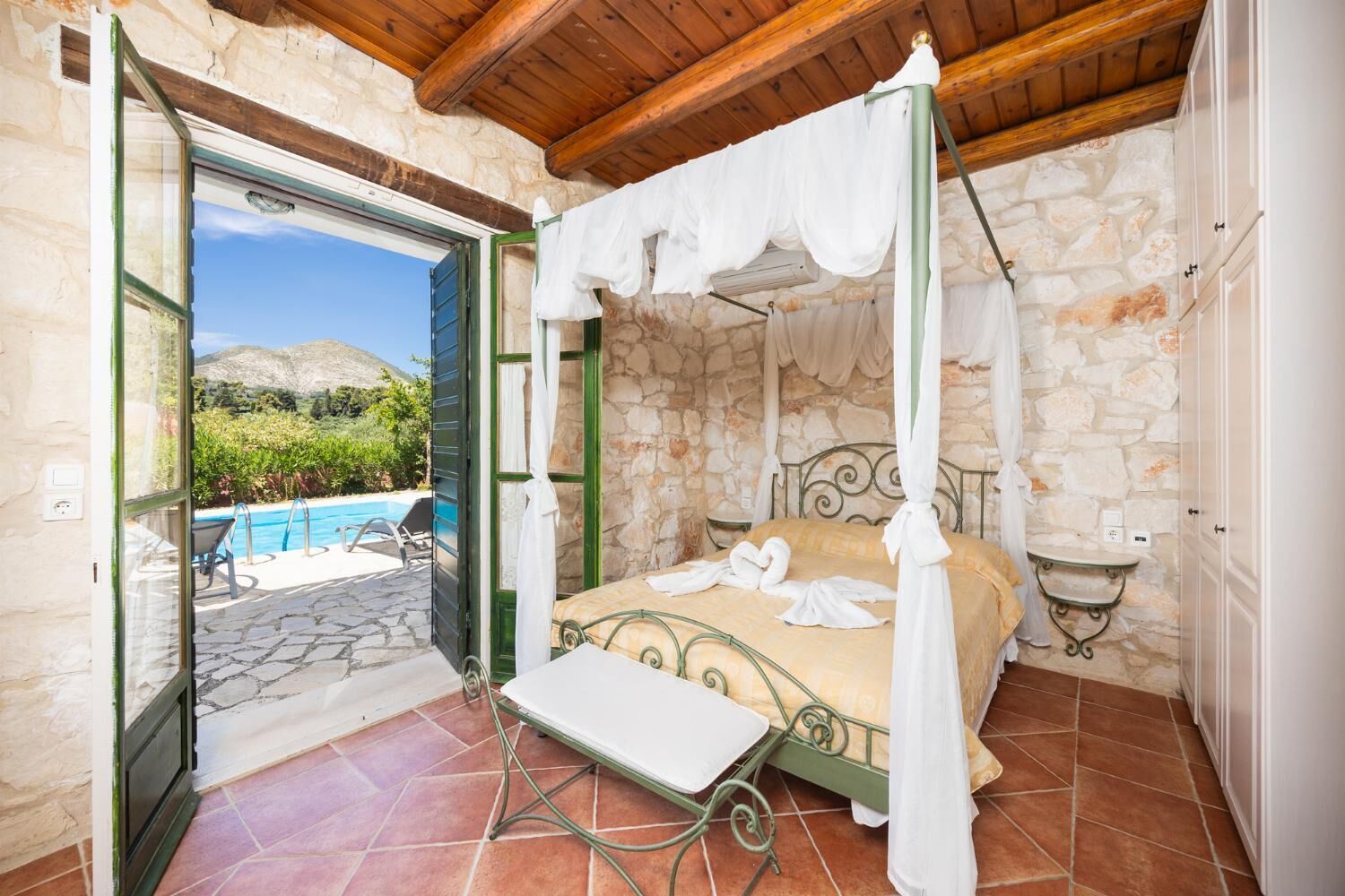 Une chambre avec un mur en pierre, un lit à baldaquin, une petite table et une vue sur une piscine et des montagnes.