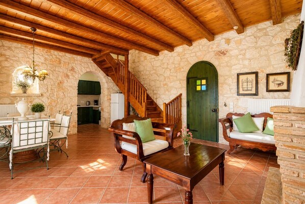 Un interior rústico con una escalera de madera, paredes de piedra, un comedor con mesa y sillas, y una puerta verde.