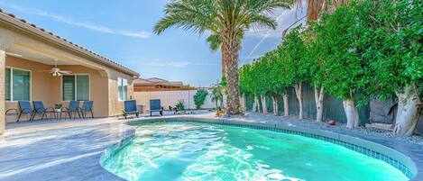 A swimming pool with a curved edge, surrounded by palm trees and a patio area with chairs and a ceiling fan.