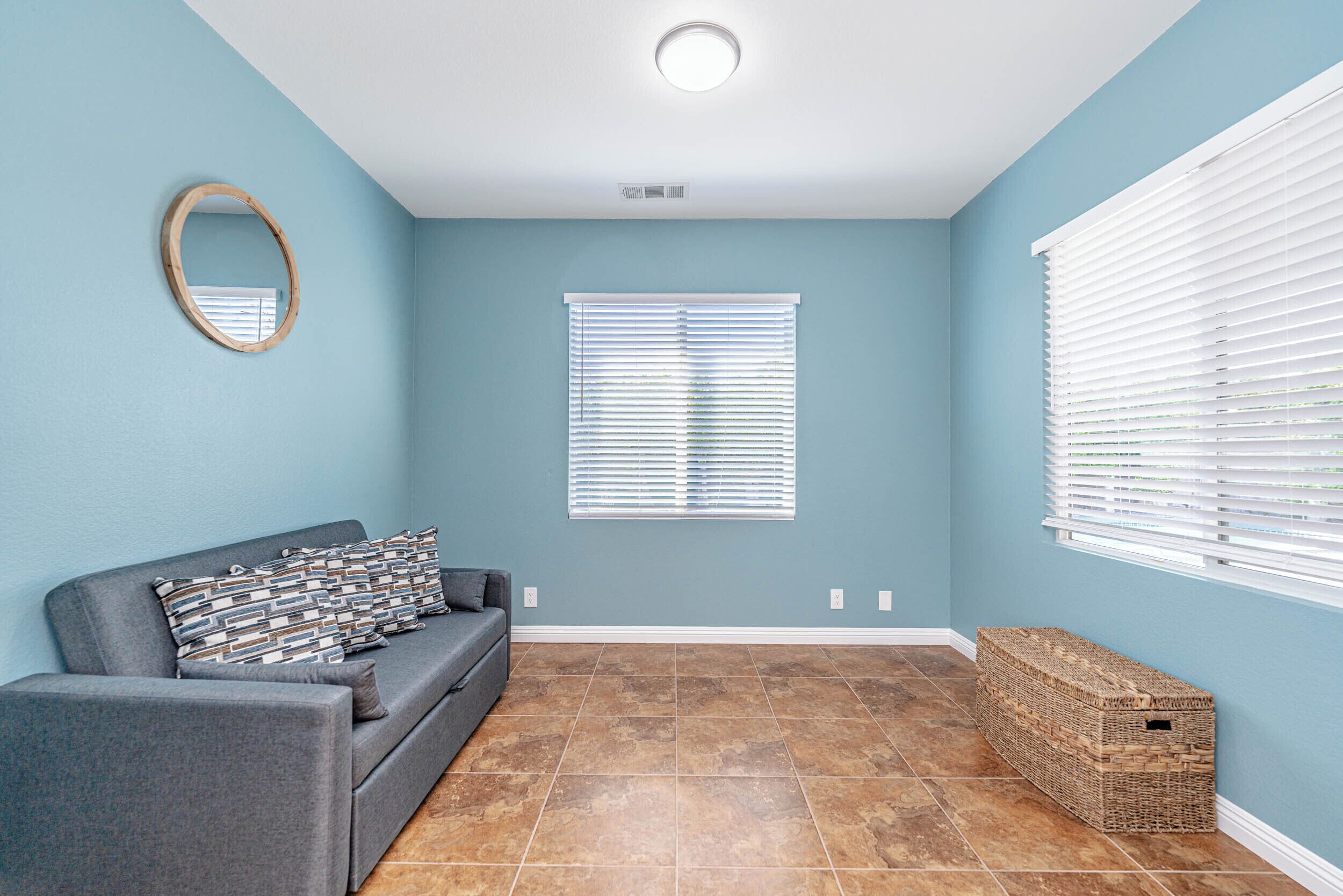 A room with a grey sofa, a wooden-framed mirror, a window with blinds, and a wicker basket.