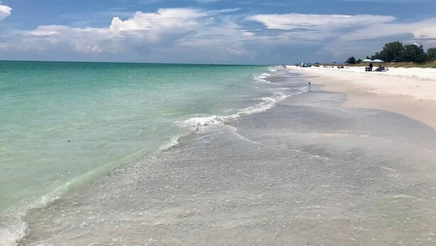 A beach with clear turquoise water, a sandy shore, and a few structures in the distance.