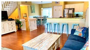 A spacious living room with a wooden coffee table, a blue sofa with colorful cushions, and a kitchen area with white cabinets and blue bar stools.