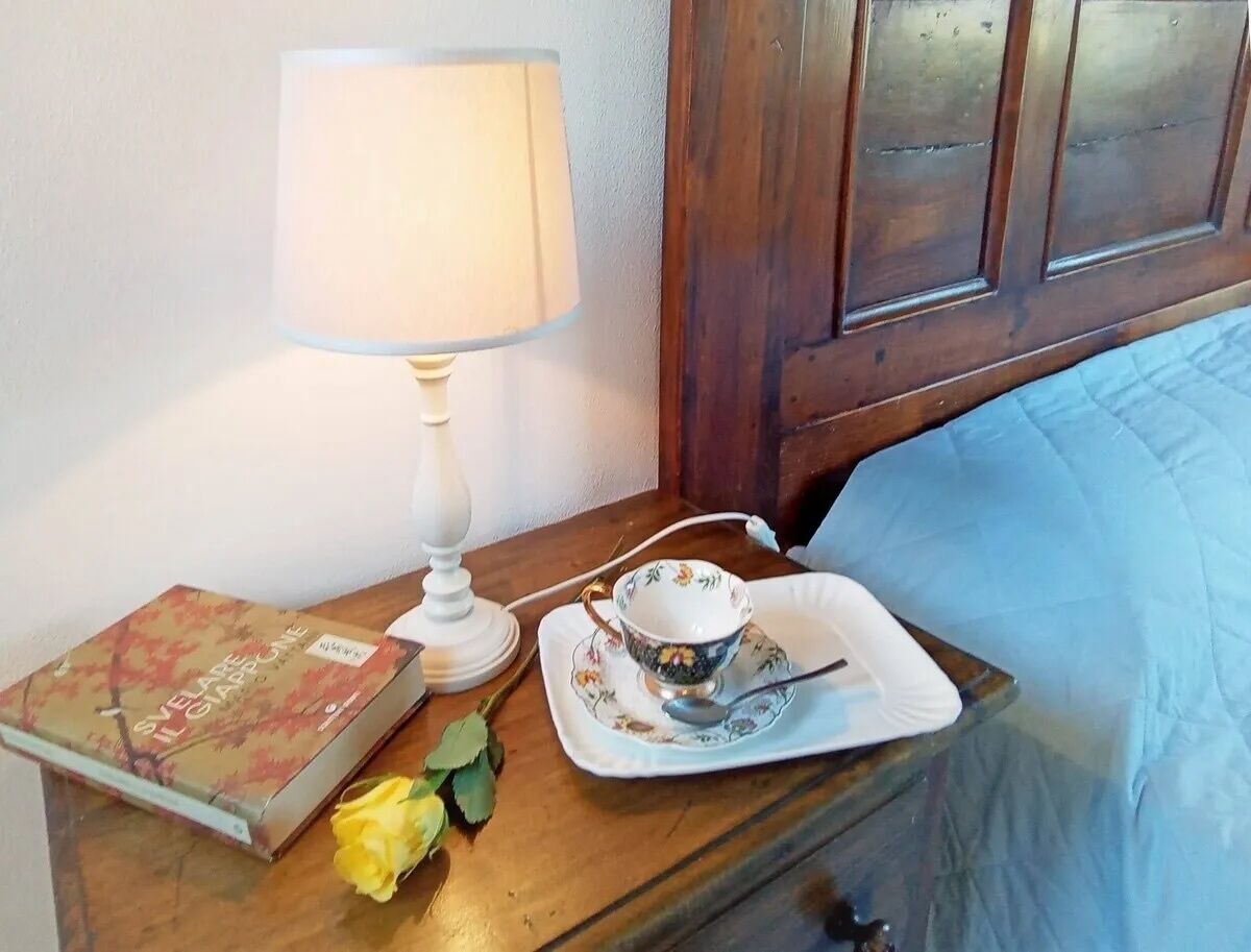 Een nachtkastje met een boek, een theekopje, een schoteltje, een lamp en een enkele gele roos.