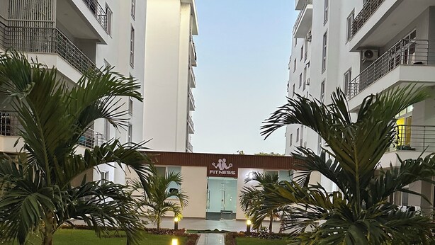 Un vialetto conduce a un edificio con un cartello 'FITNESS', affiancato da palme e prato curato.