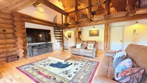 A log cabin interior with a wooden staircase, a sofa, a television, and a patterned rug. - VT Lodge Cabin w Mountain Views near Killington (Proctor)