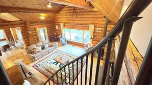 A log cabin interior with a wooden staircase, a living room with a sofa and chairs, a dining area with a table and chairs, and large windows. - VT Lodge Cabin w Mountain Views near Killington (Proctor)