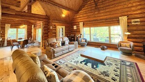 A log cabin interior with a cozy living room, wooden ceiling beams, and large windows. - VT Lodge Cabin w Mountain Views near Killington (Proctor)