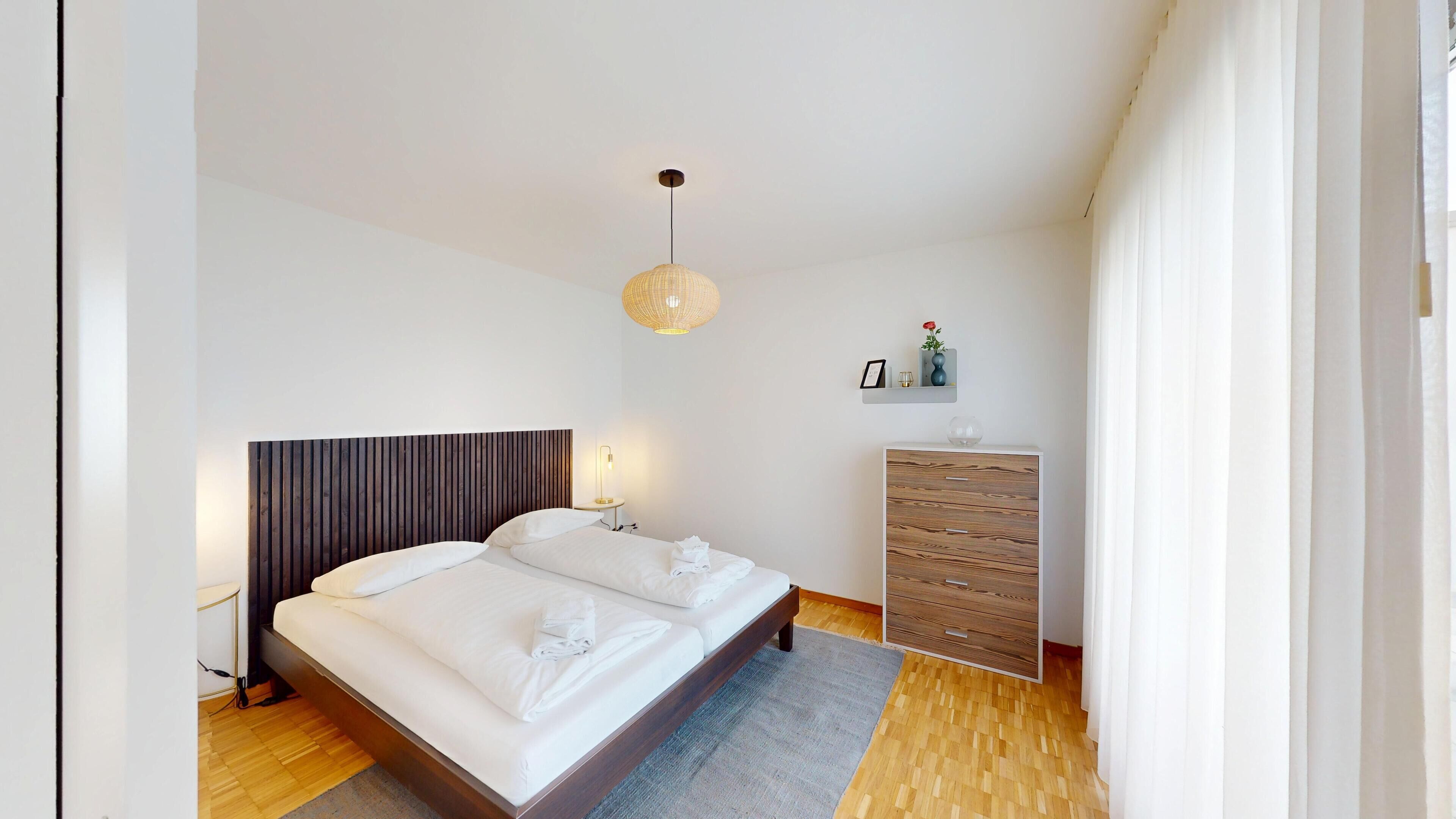 A modern bedroom with a large bed, bedside lamps, a wooden headboard, a bedside table, a wardrobe, and a hanging light fixture.