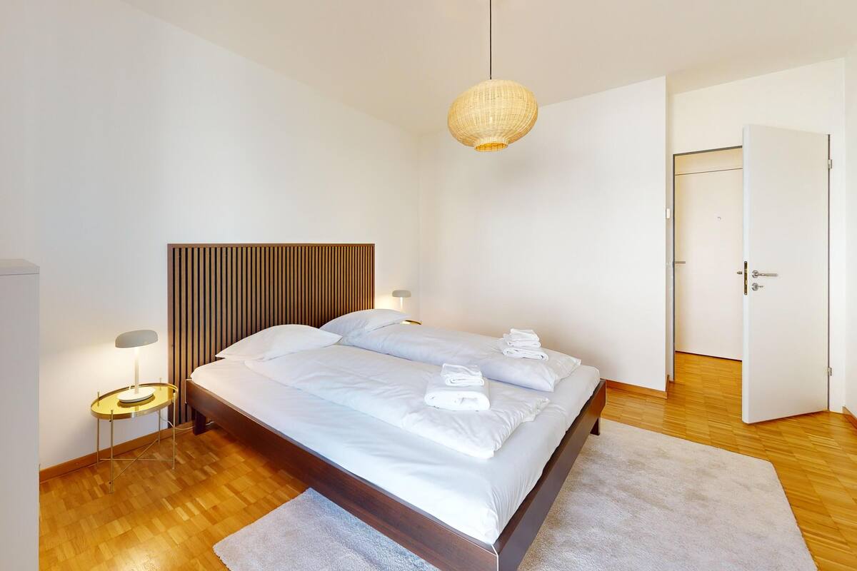 A modern bedroom with a wooden headboard, a large bed with white linens, two bedside lamps, and a hanging light fixture.