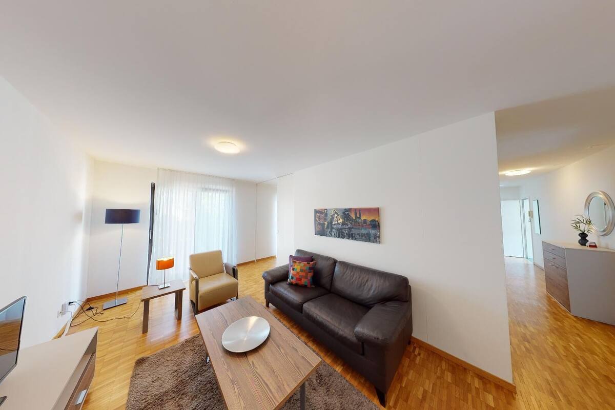 A modern living room with a sofa, a coffee table, and a TV. There is a dining area with a table and chairs. The room has wooden flooring and white walls.