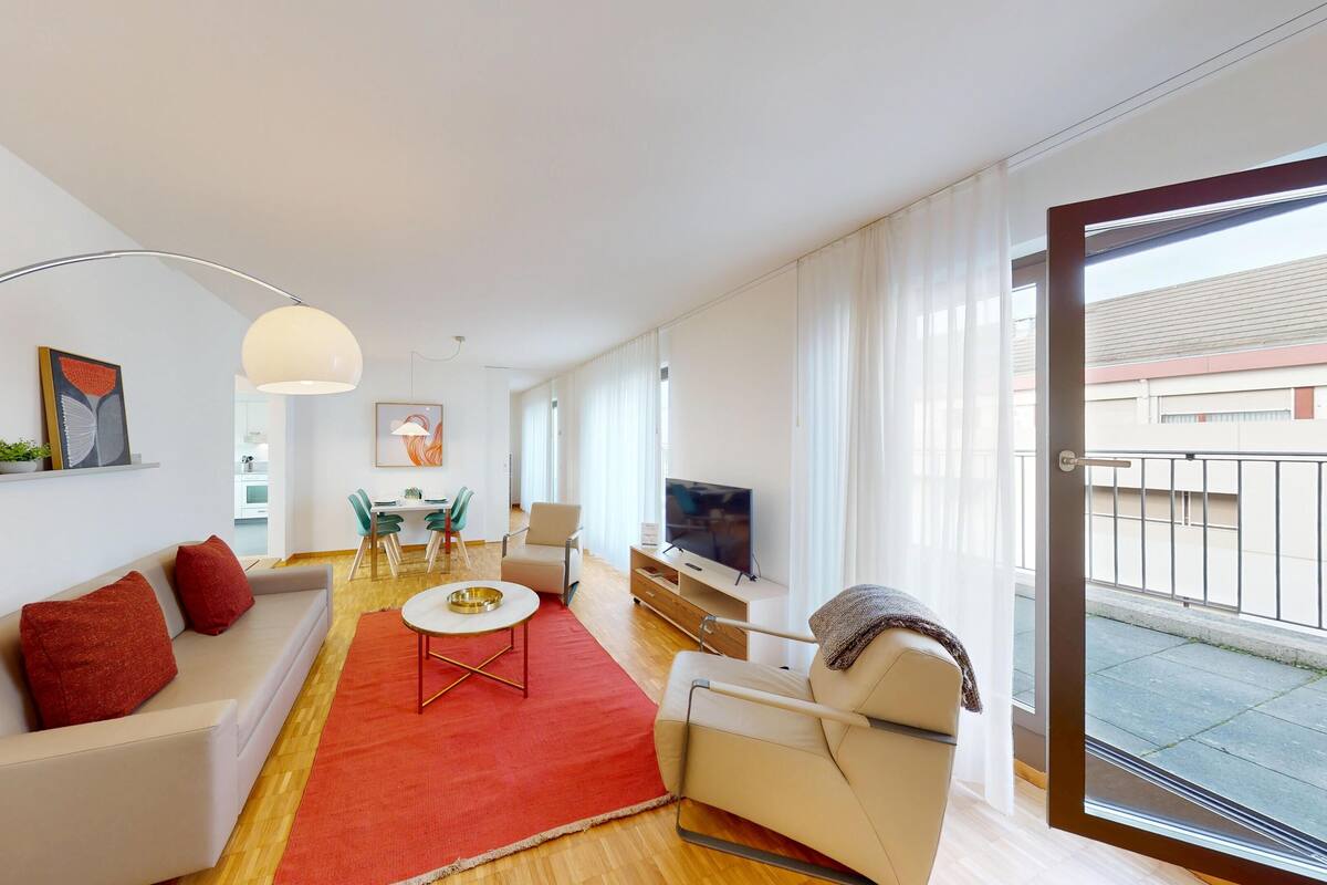 A modern living room with a sofa, armchair, and TV, a dining area with chairs, and a large window with a view.