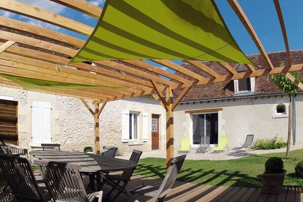 An outdoor patio area with a wooden pergola, a round table with chairs, and a lawn with a few lounge chairs.