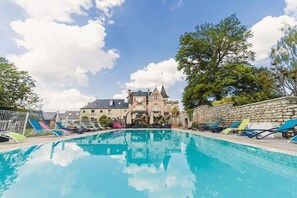 A swimming pool with lounge chairs and a stone wall, surrounded by trees and a large building with a tower.