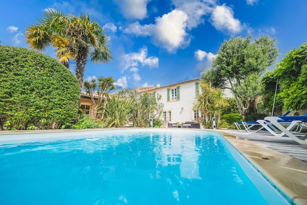A swimming pool with lounge chairs, surrounded by lush greenery and a white building with a tiled roof.