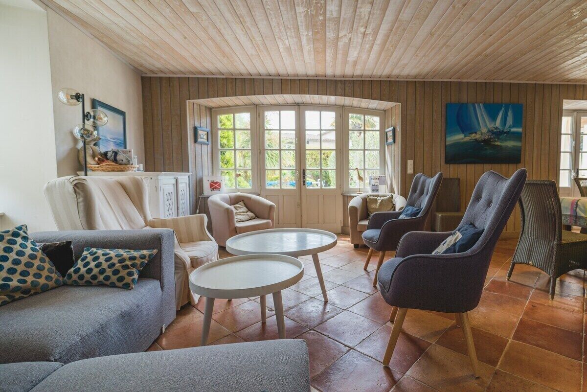 Een gezellige woonkamer met een houten plafond, grote ramen en comfortabele zitplaatsen, waaronder een bank en fauteuils.