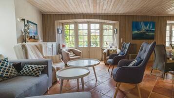 Een gezellige woonkamer met een houten plafond, grote ramen en comfortabele zitplaatsen, waaronder een bank en fauteuils.