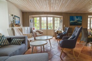 Ein gemütliches Wohnzimmer mit Holzbalkendecke, großen Fenstern und bequemen Sitzgelegenheiten, darunter eine Couch und Sessel.