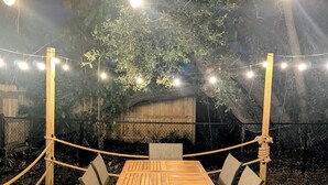 Una terraza de madera al aire libre con una mesa y sillas, rodeada por una valla de cuerda, bajo luces colgantes.