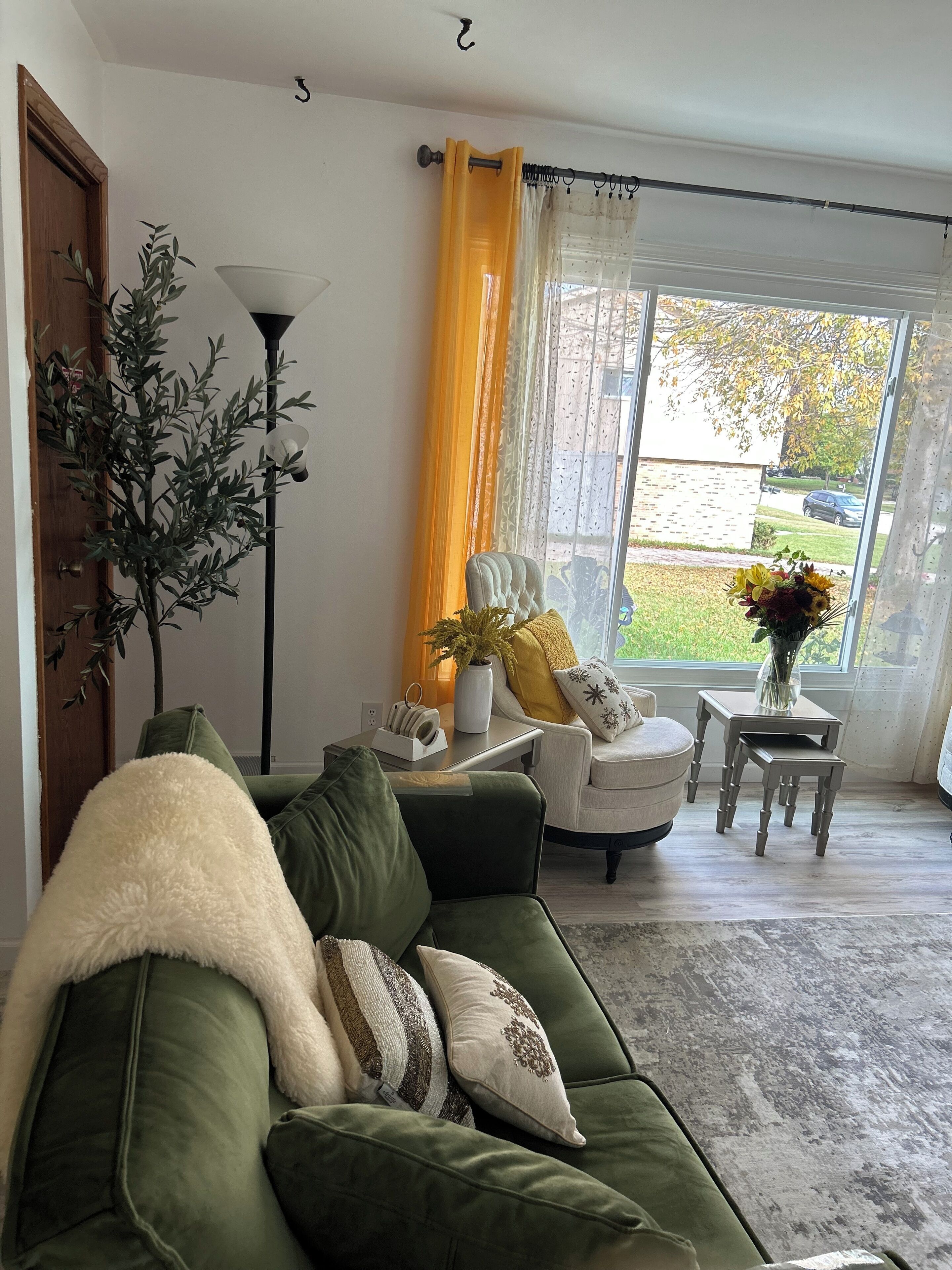 Ett vardagsrum med en grön soffa, en krukväxt, en lampa, en stol, ett litet bord med en vas och ett fönster med gardiner.