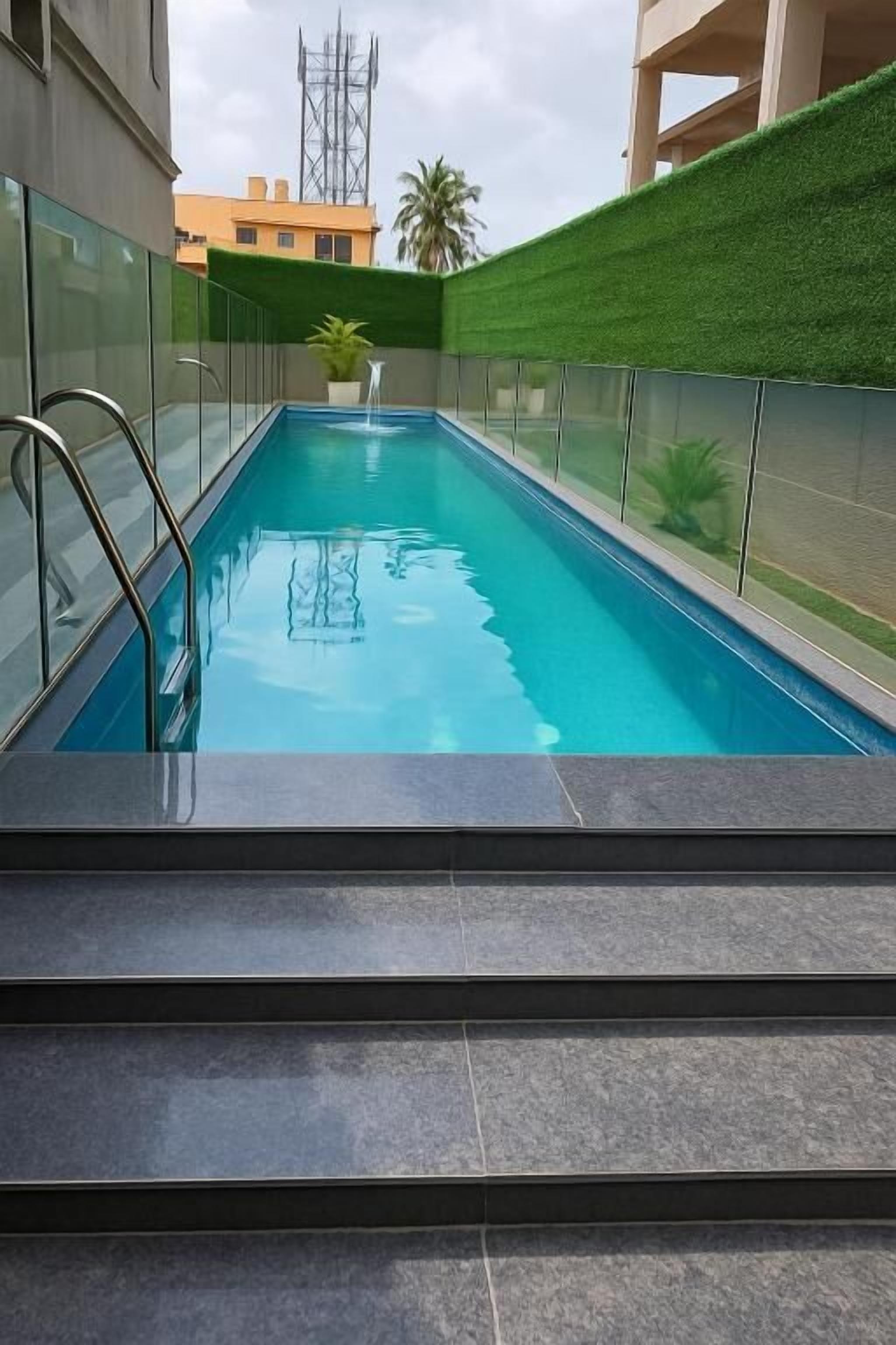 A rectangular indoor swimming pool with clear blue water, surrounded by glass railings and a green artificial turf wall.