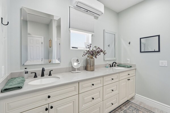 A bathroom with two sinks, a mirror, a vase with flowers, and a window with blinds.