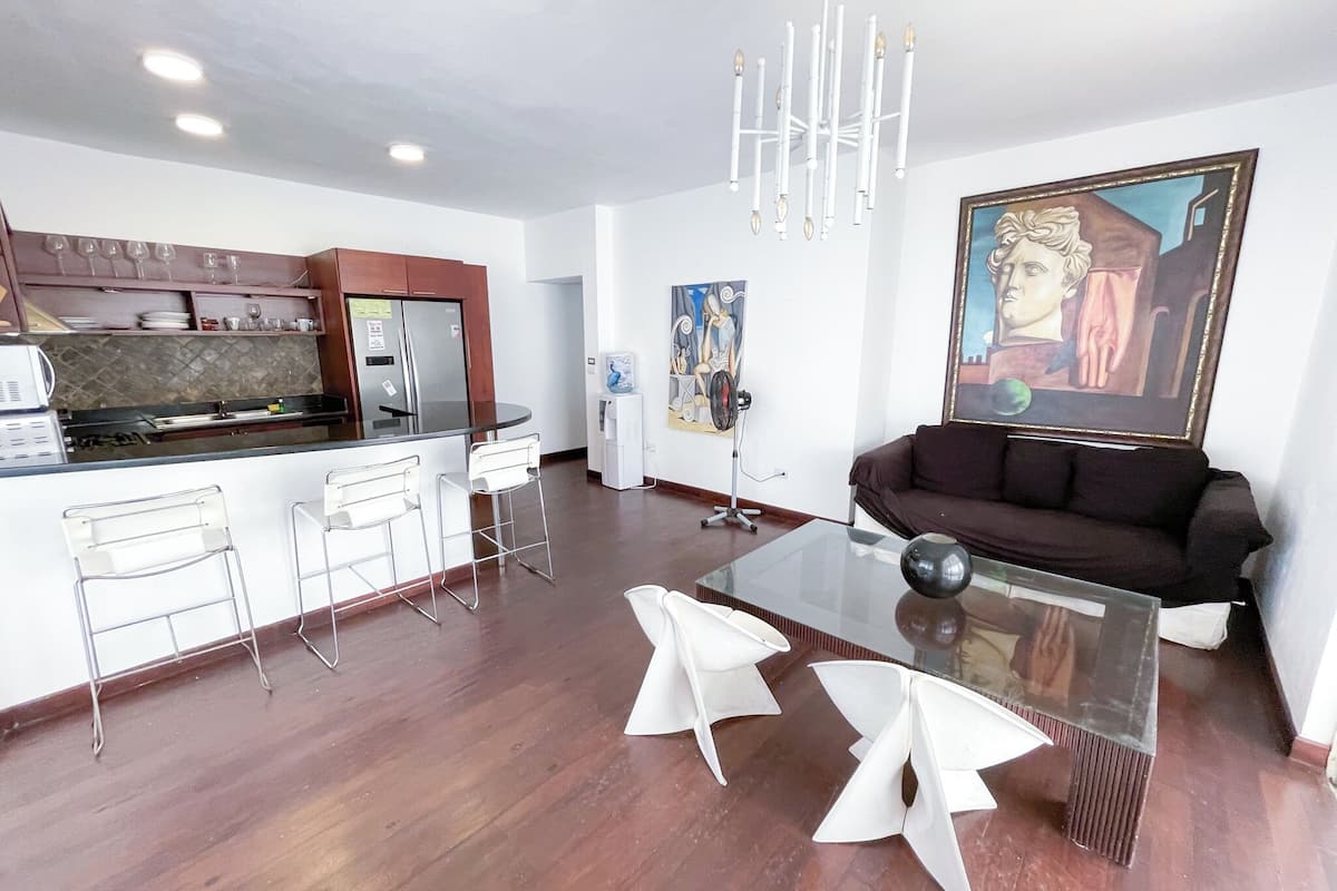 Una sala de estar moderna con una mesa de centro de vidrio, sillas blancas, un sofá oscuro y un cuadro en la pared.