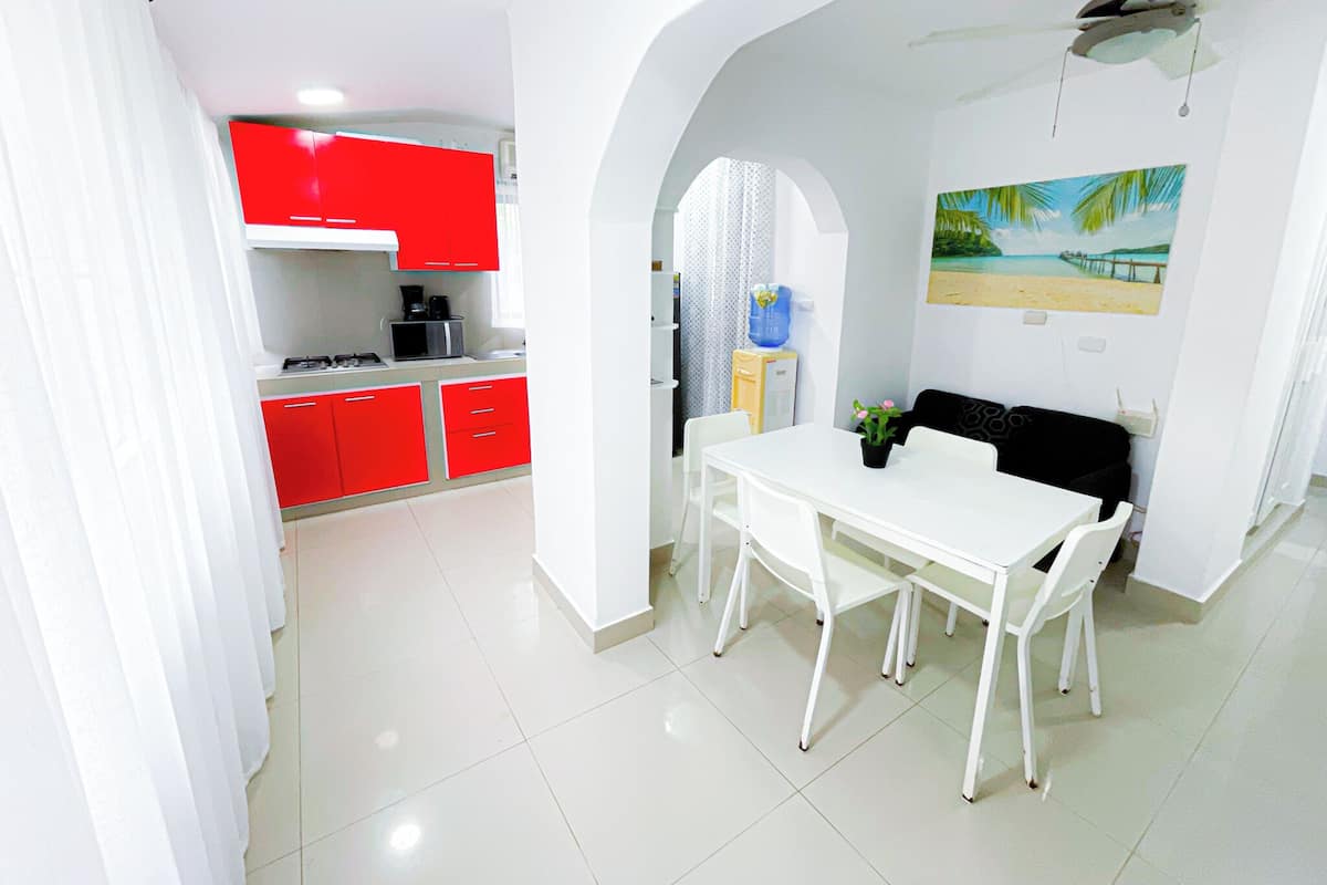 Una cocina moderna con armarios rojos, una zona de comedor blanca con mesa y sillas, y un cuadro de una escena de playa en la pared.