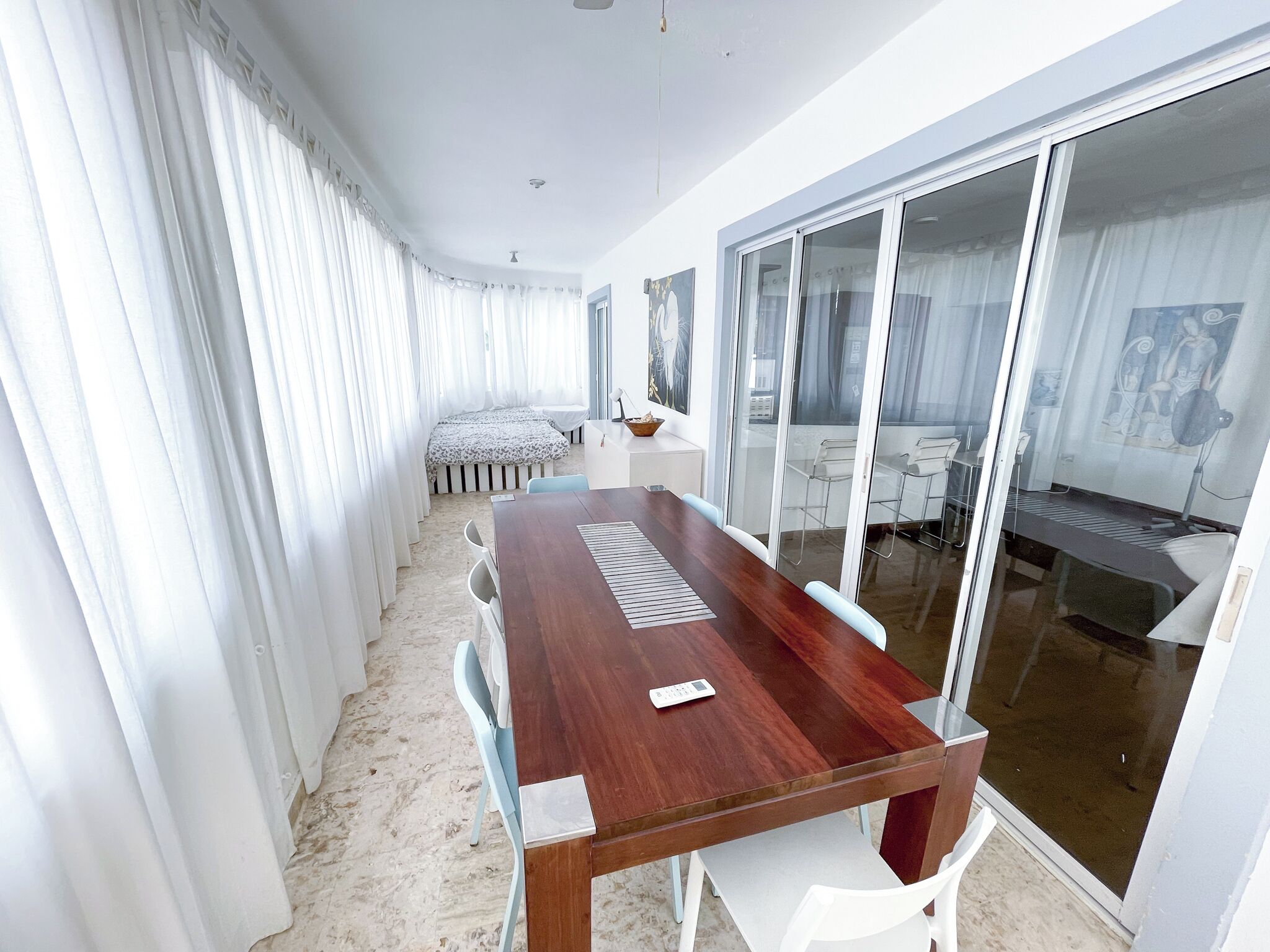 A modern hotel room with a large wooden dining table, white chairs, a bed with a striped headboard, and a glass door leading to a balcony with a view.