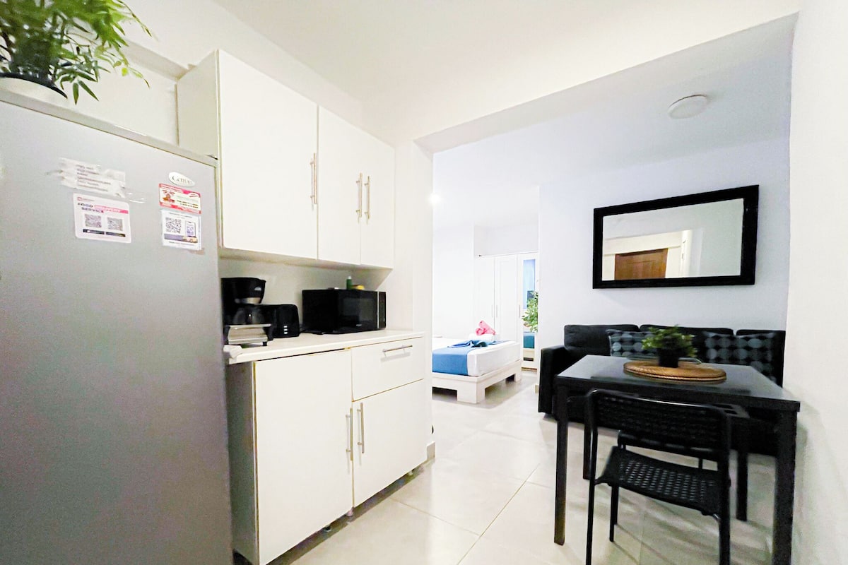 Cocina pequeña con gabinetes blancos, un refrigerador, un microondas y una cafetera. Hay un comedor con una mesa y sillas, y una cama al fondo.