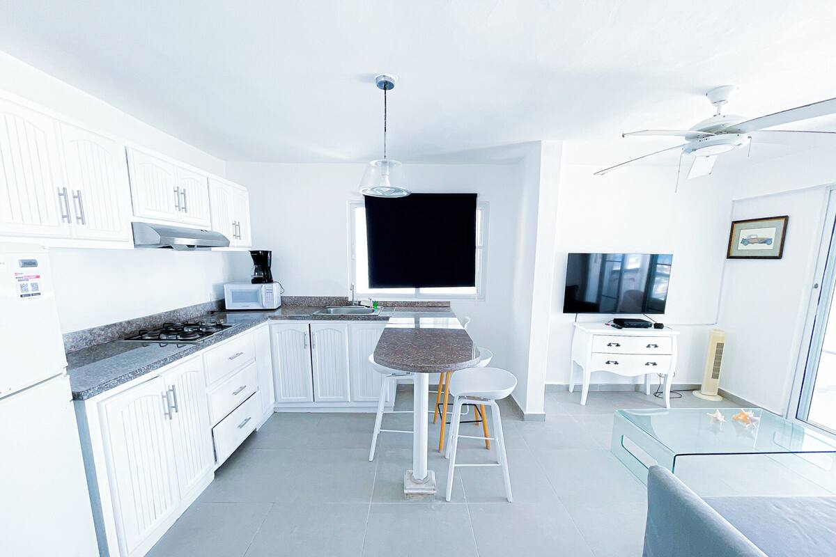 Una cocina moderna con armarios blancos, una estufa a gas, un microondas y una zona de comedor con taburetes.