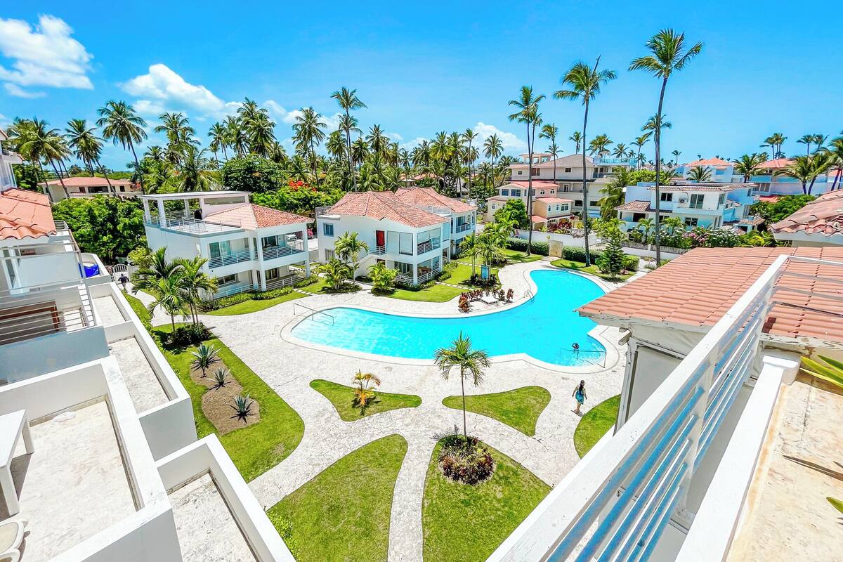 Un resort con piscina central, varias edificaciones con techos de tejas rojas y palmeras.