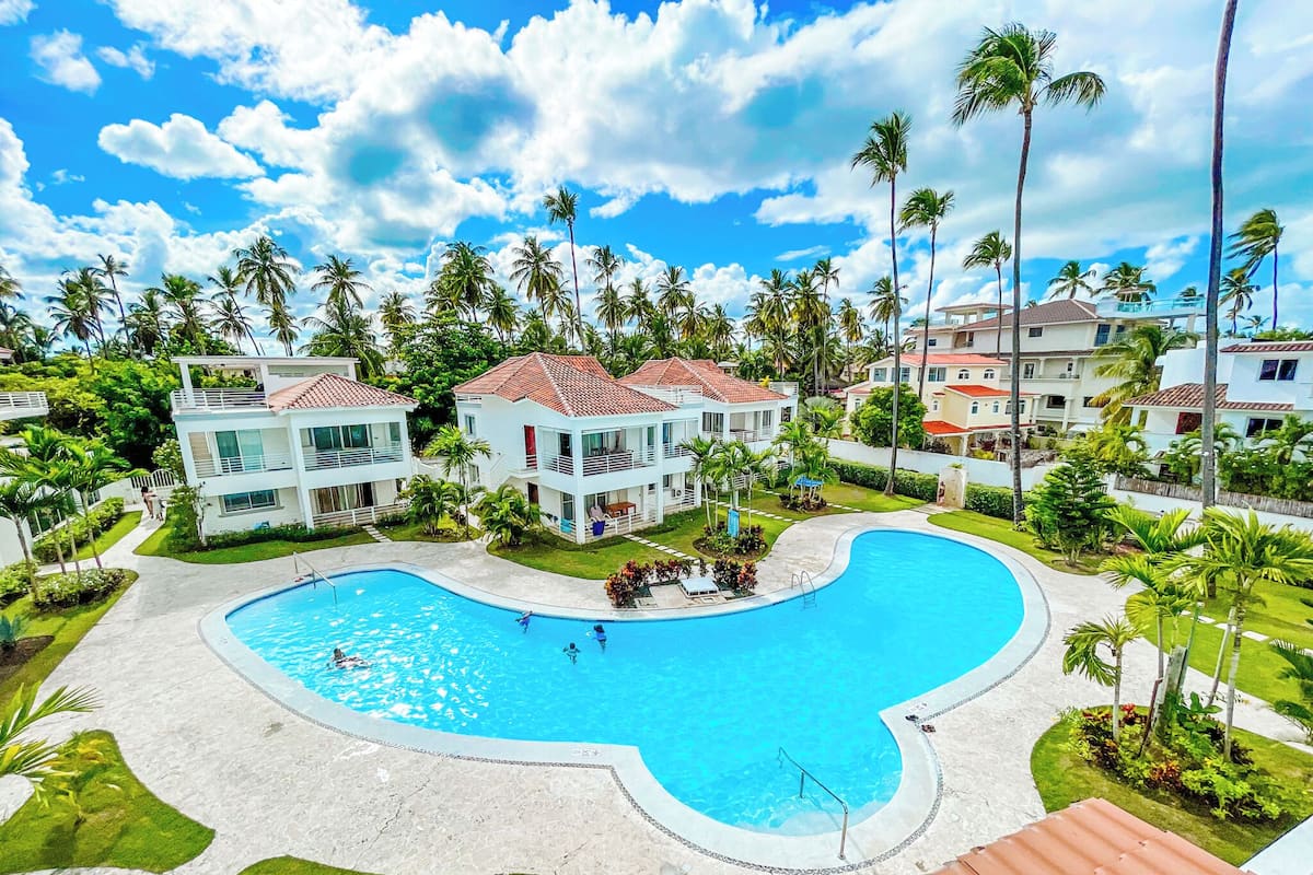 Un resort con una amplia piscina, varias edificaciones de dos pisos y palmeras.