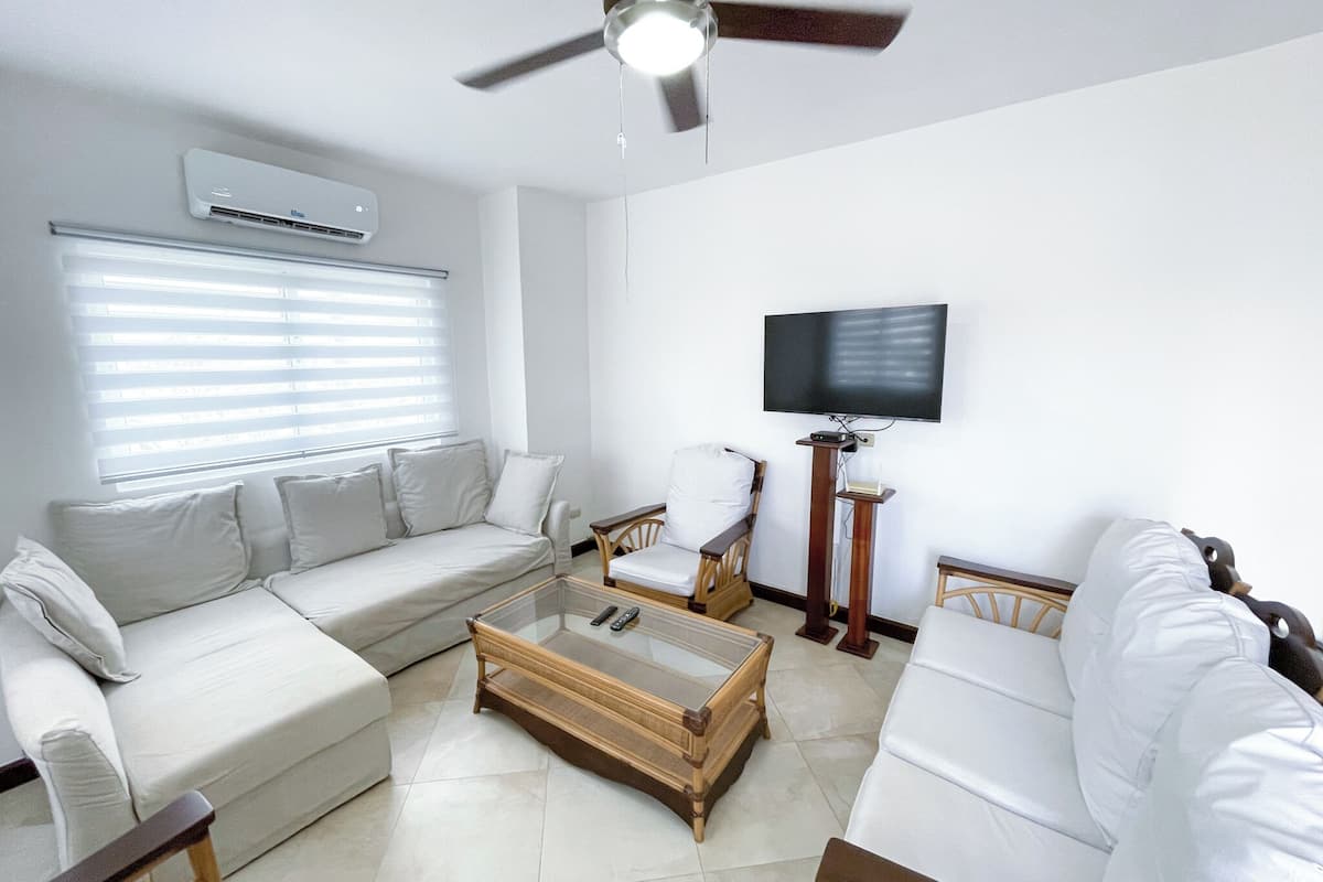 Una sala de estar con un sofá blanco, una mesa de centro de cristal, una silla de madera, un televisor montado en la pared y un ventilador de techo.
