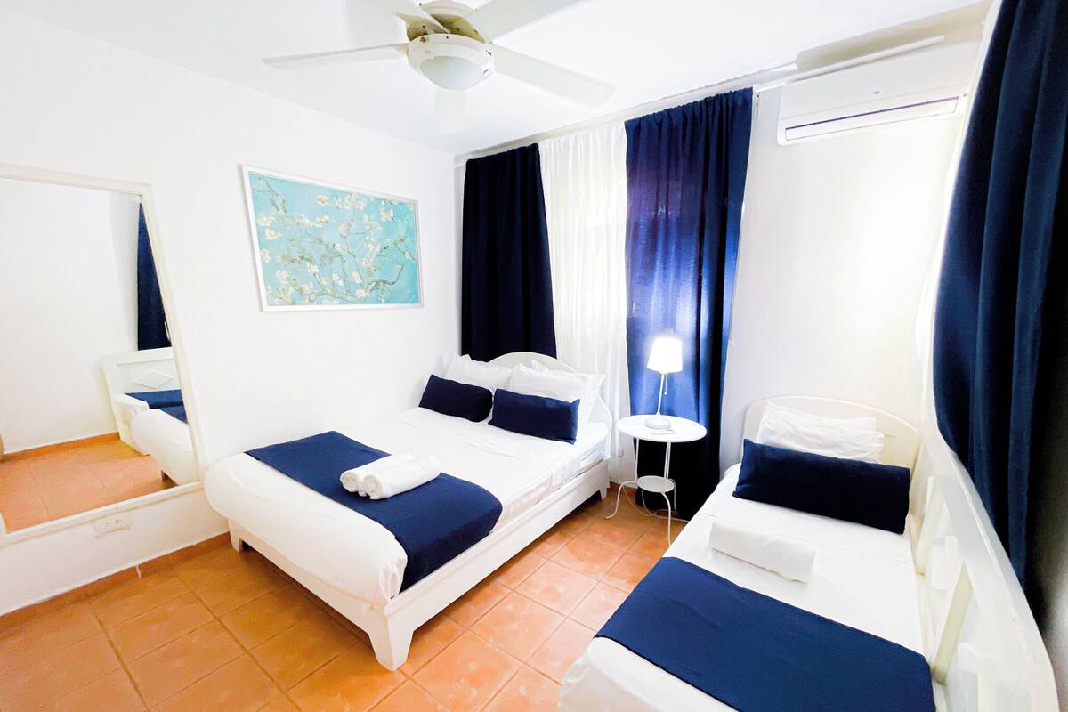 Habitación de hotel con dos camas, ventilador de techo, una mesita con lámpara y un cuadro en la pared.