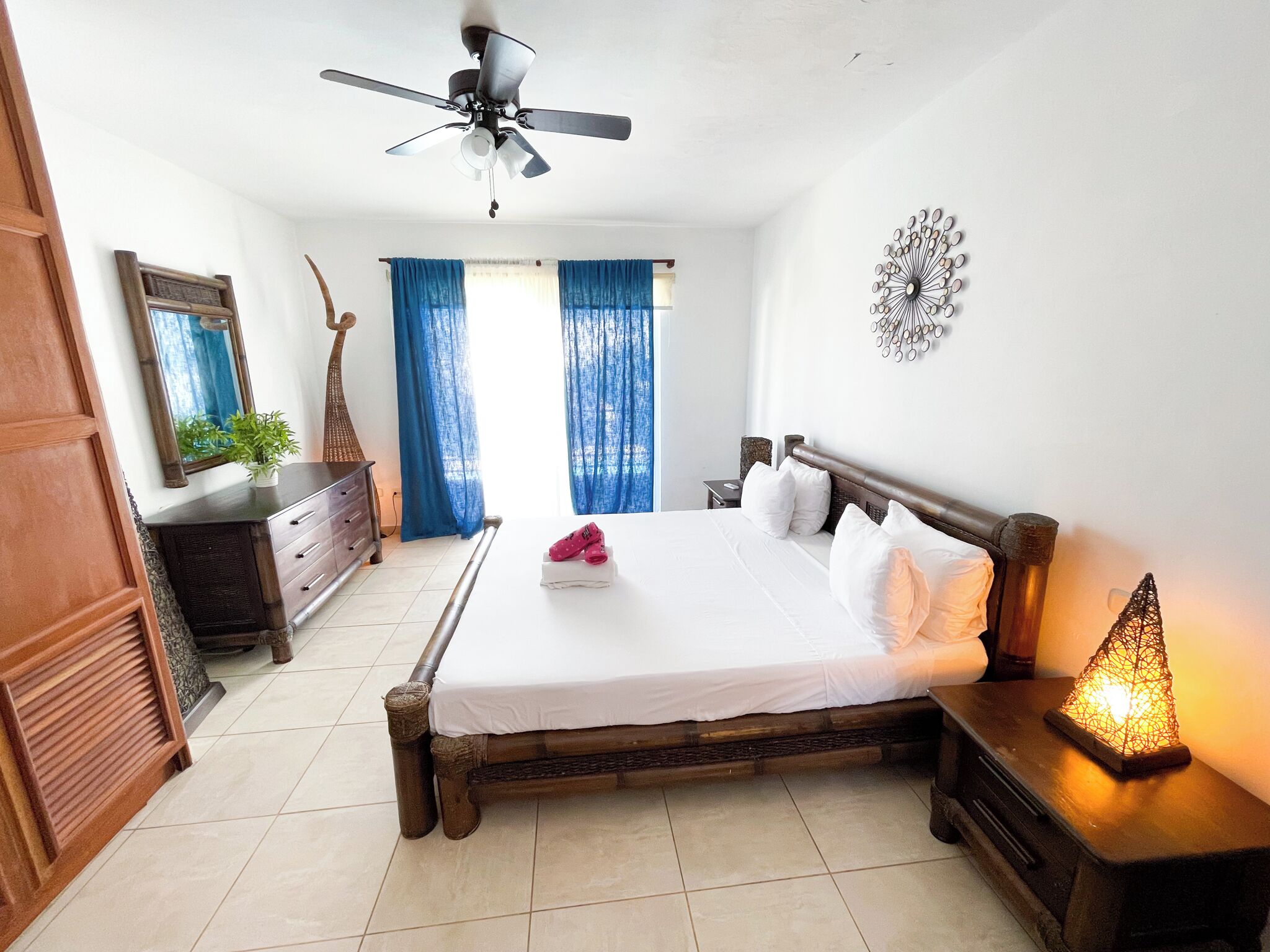 A bedroom with a wooden bed, a ceiling fan, a nightstand with a lamp, a dresser, and a large window with blue curtains.