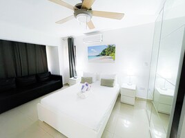 A modern hotel room with a large bed, a ceiling fan, a nightstand, a lamp, and a window with a beach view.