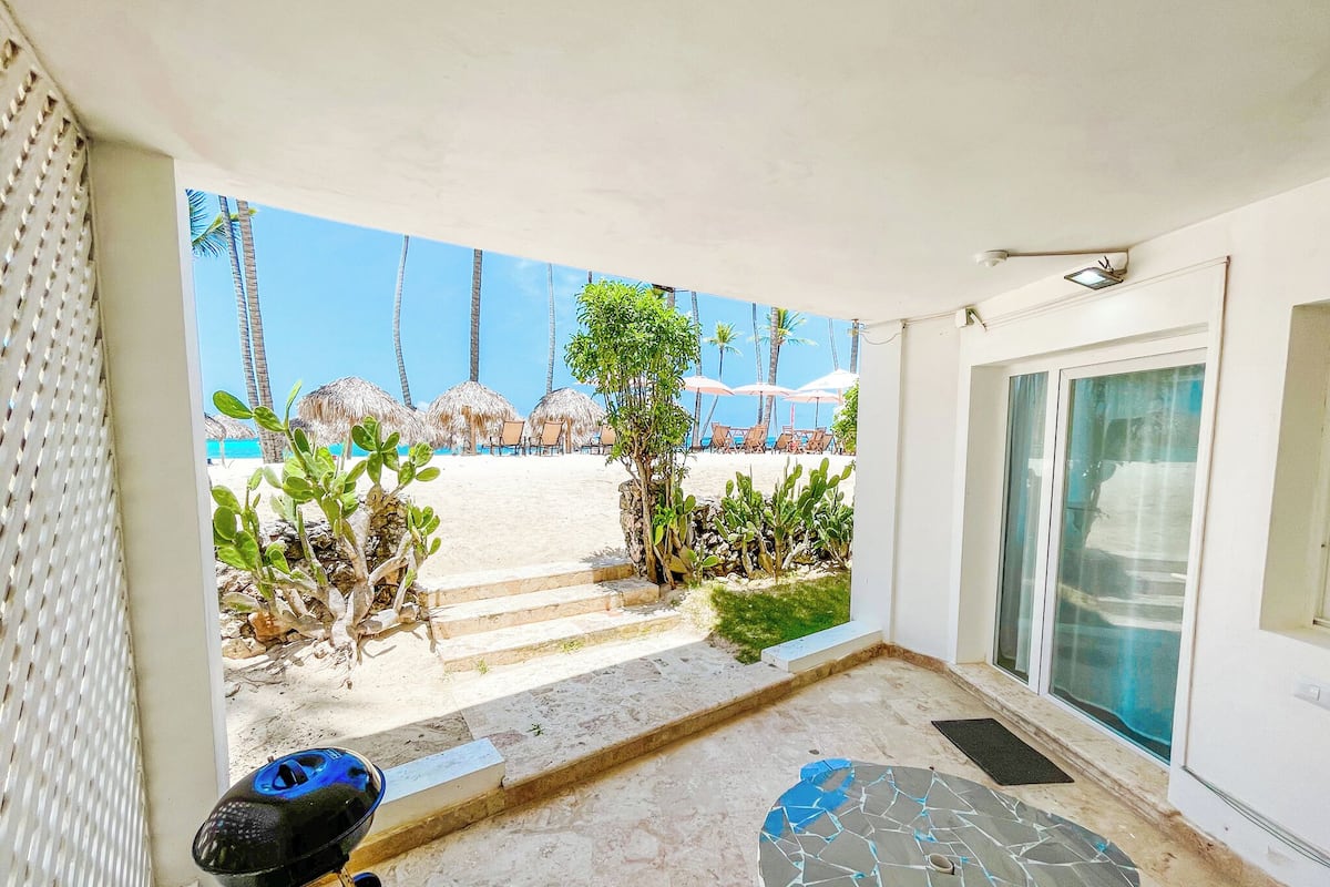 Un patio con una parrilla, una mesa de mosaico y vistas a una playa con sombrillas y sillas.