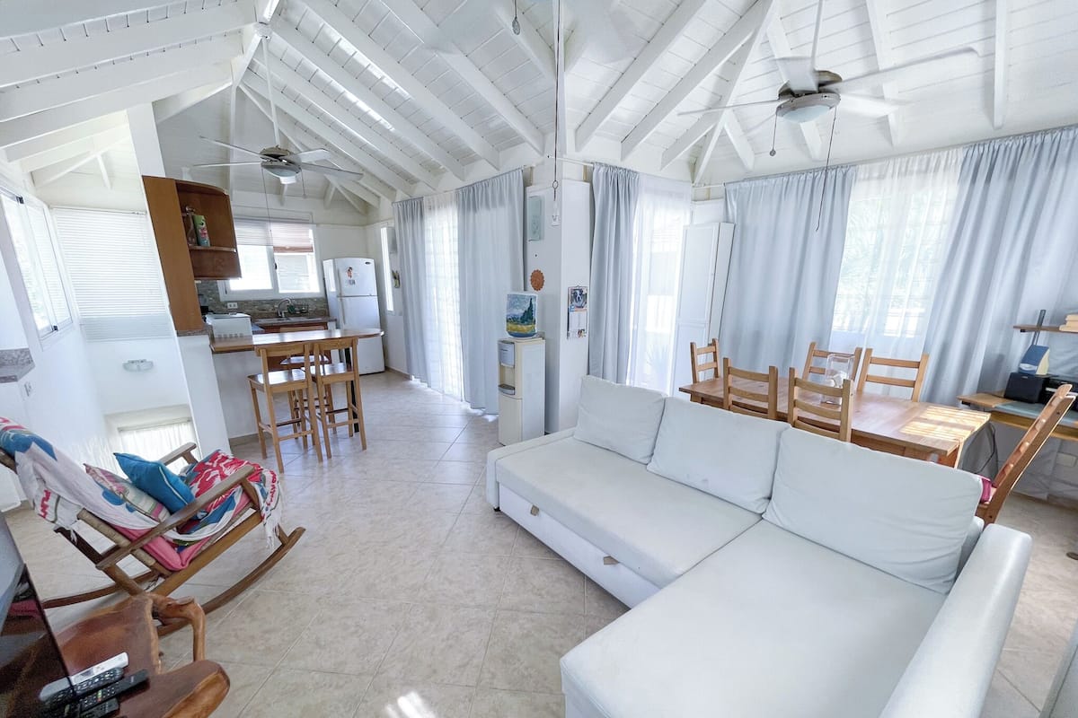 Amplia sala de estar con un sofá blanco, mesa de comedor de madera y sillas. Se ve un área de cocina con un refrigerador y una mesa de comedor al fondo.