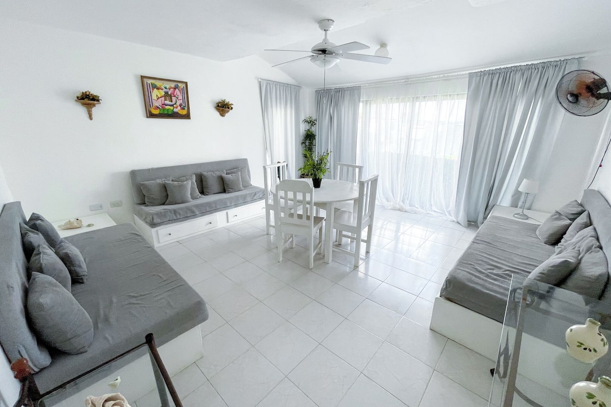 Una habitación moderna y bien iluminada, con un sofá gris, una mesa de comedor con sillas, un ventilador de techo y un ventanal con cortinas translúcidas.