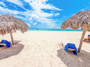 A beach with thatched umbrellas and lounge chairs, a clear sky, and the ocean in the background.