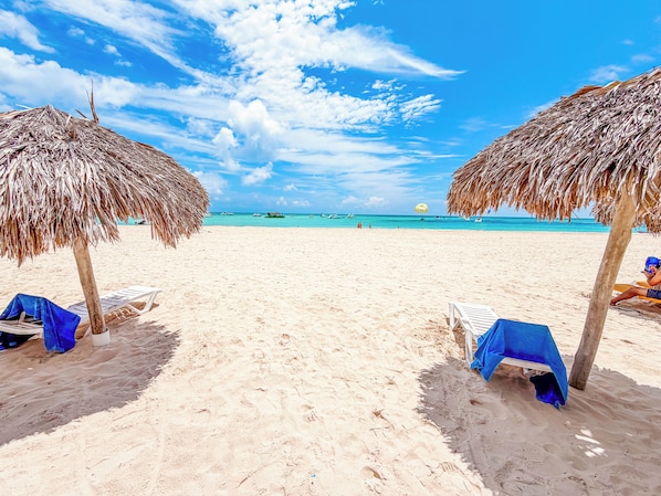 A beach with thatched umbrellas and lounge chairs, a clear sky, and the ocean in the background.