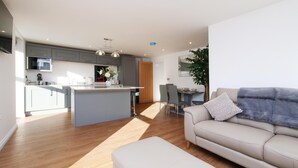 Eine moderne Küche mit grauen Schränken, einer zentralen Kochinsel und Pendelleuchten. Es gibt eine Essecke mit Tisch und Stühlen sowie ein Wohnzimmer mit einer Couch und einer Pflanze.