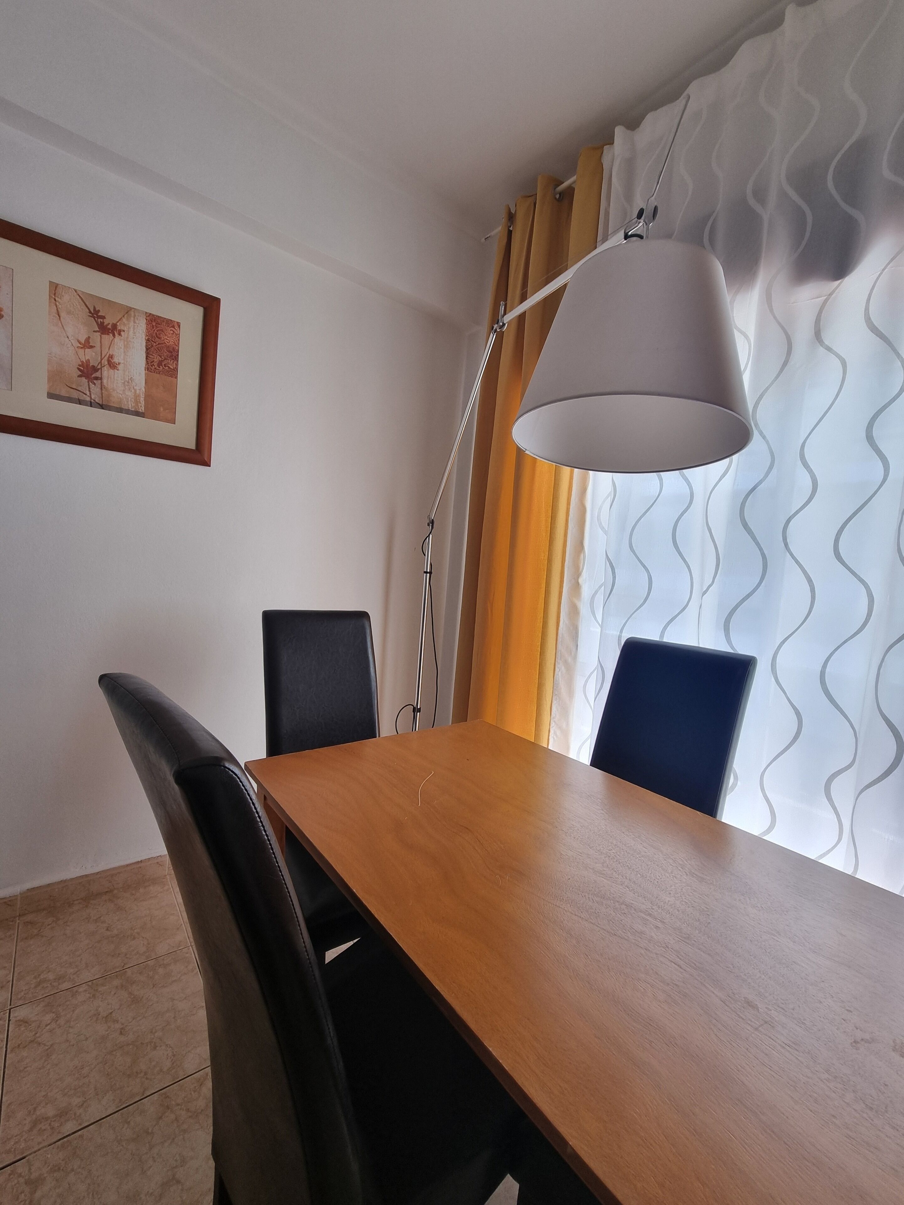 A dining area with a wooden table, black chairs, a floor lamp, and a framed picture on the wall.