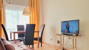Ein Wohnzimmer mit einem braunen Ledersofa, einem Esstisch mit Stühlen, einem Fernseher auf einem goldenen Ständer und einem Fenster mit gelben Vorhängen.
