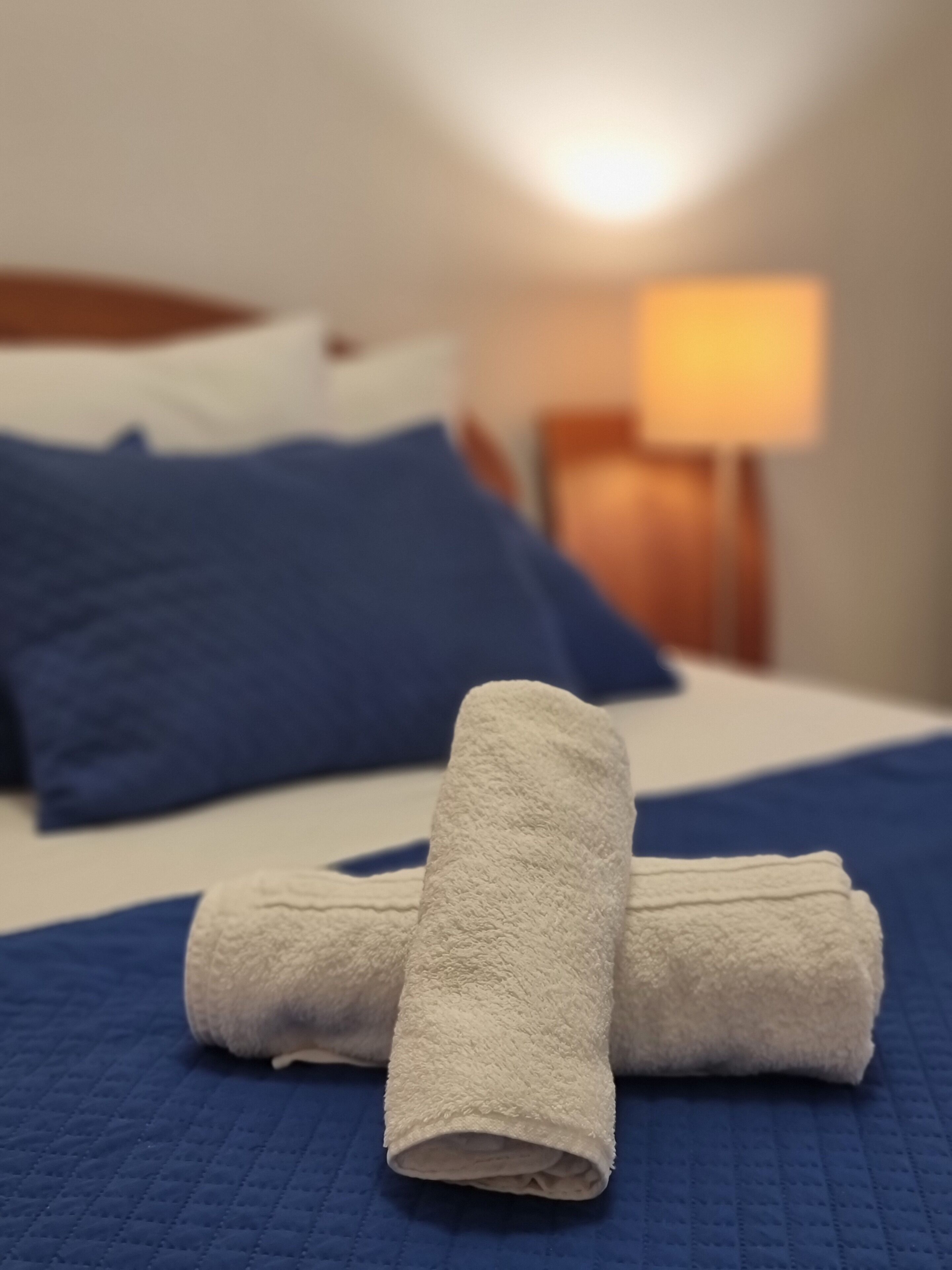 Een netjes opgemaakt bed met blauw-witte beddengoed, een witte handdoek gevouwen in het midden, en een lamp op het nachtkastje.