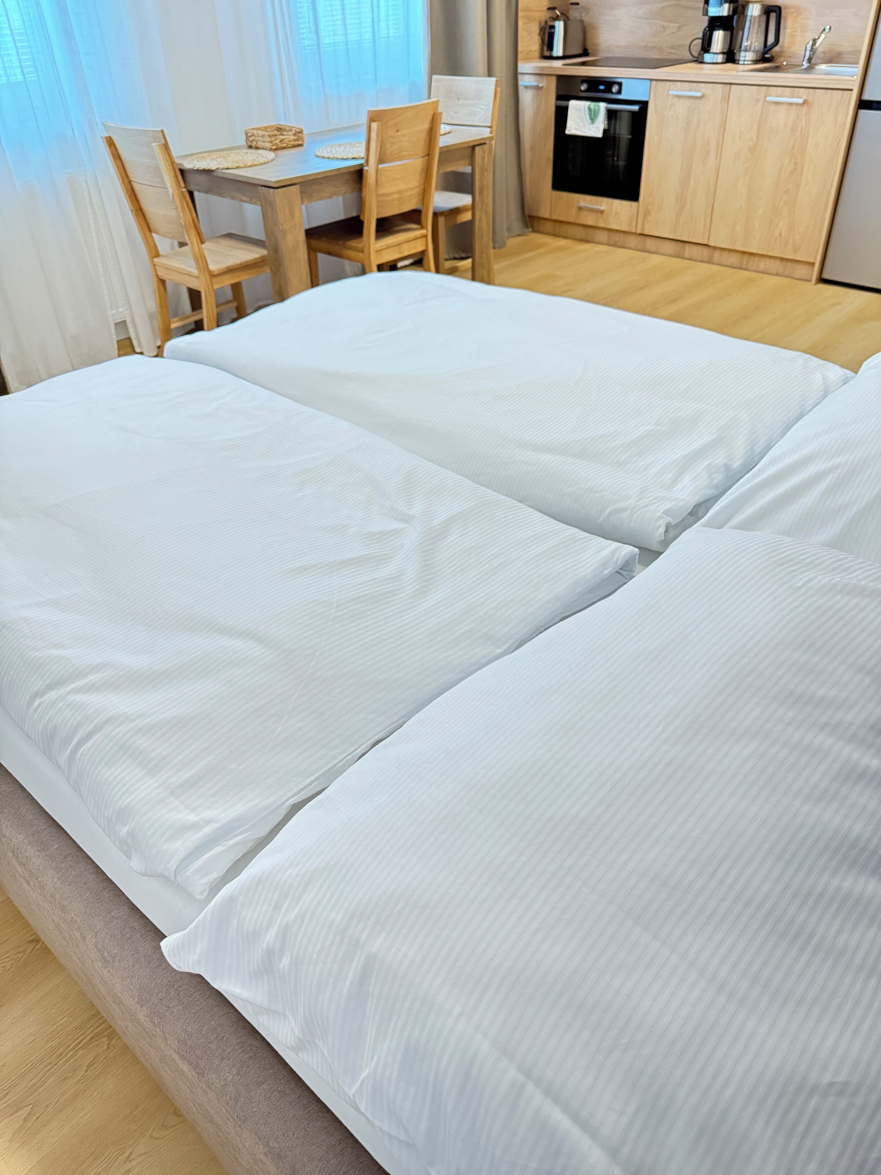 A bed with white linens in a room with a wooden dining table and chairs, a kitchen area with wooden cabinets, and a window with sheer curtains.