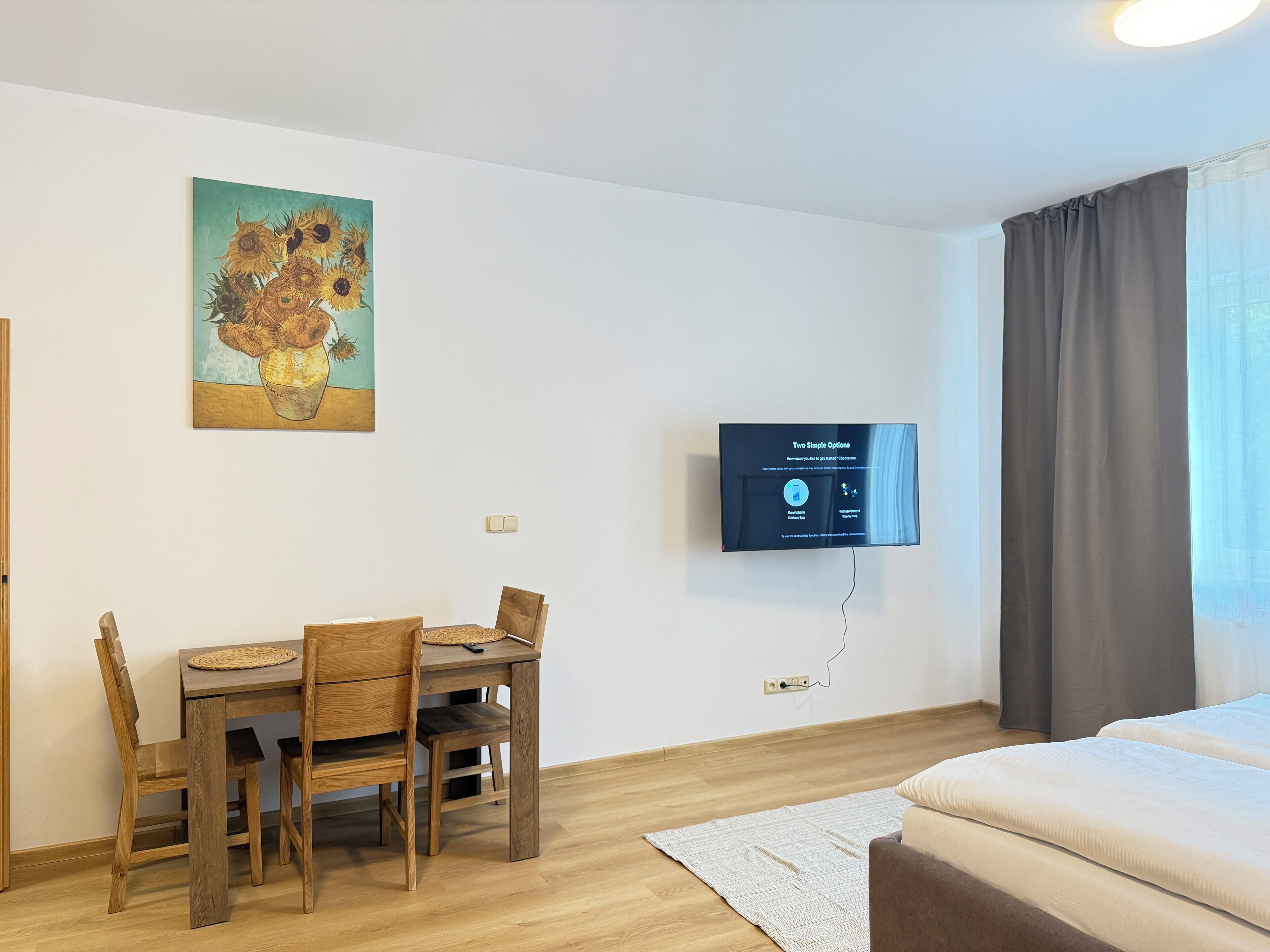 Une pièce comprenant une table à manger, des chaises, un lit, un téléviseur fixé au mur et un tableau.