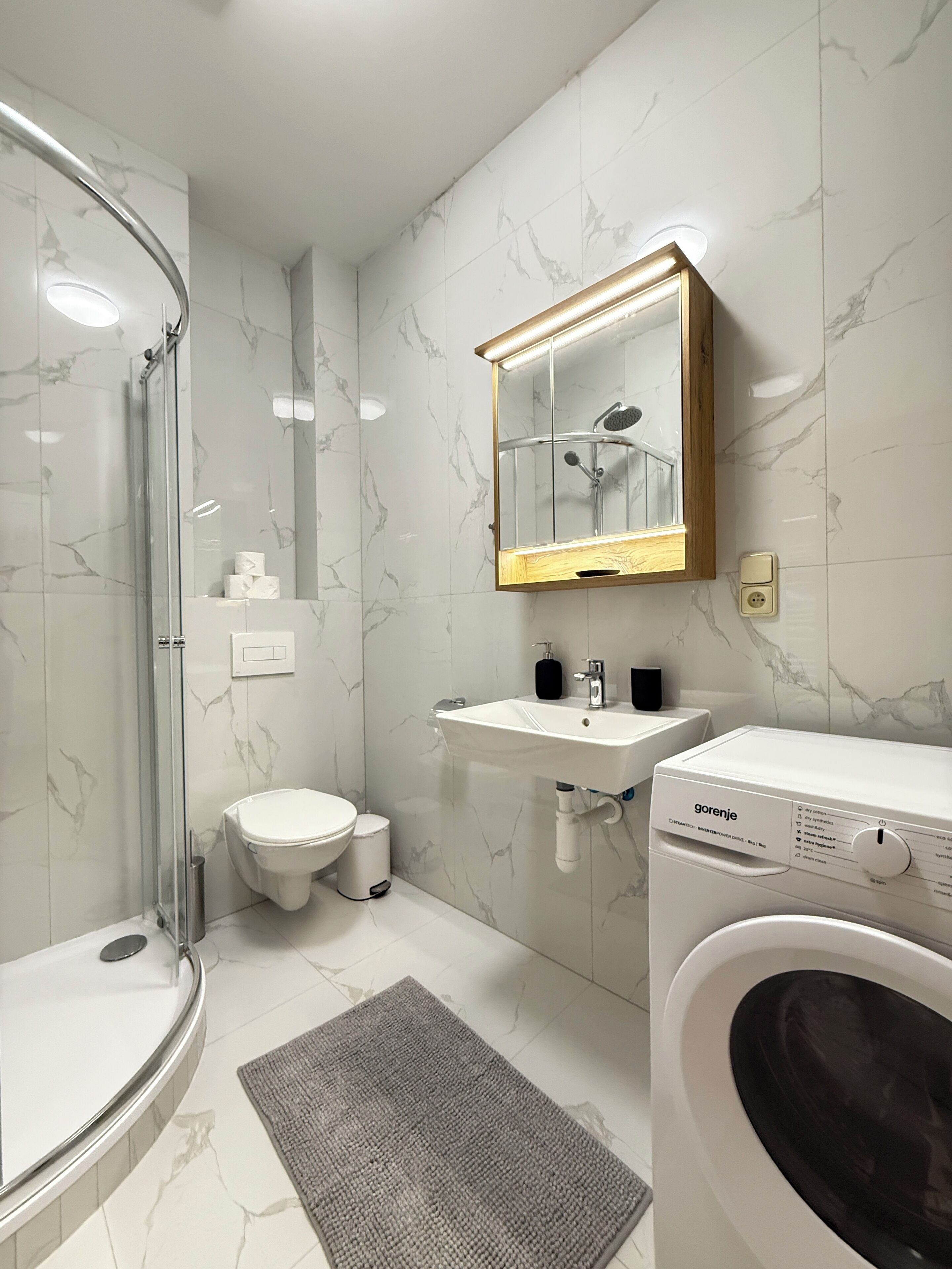 Ein modernes Badezimmer mit Duschkabine aus Glas, Toilette, Waschbecken mit Spiegel und einer Waschmaschine.