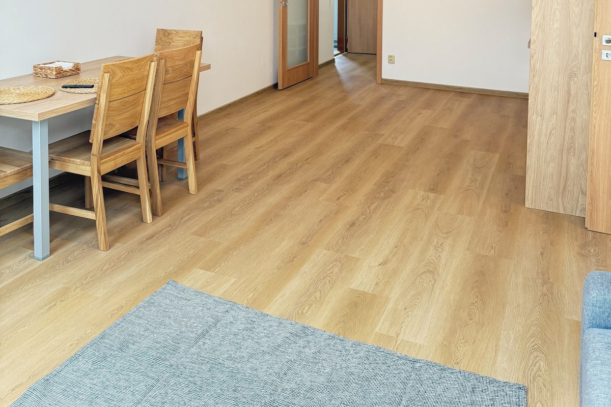 Une pièce spacieuse avec une table à manger en bois, des chaises et un tapis. On y trouve une porte qui mène à une autre pièce, une horloge au mur et des œuvres d’art.