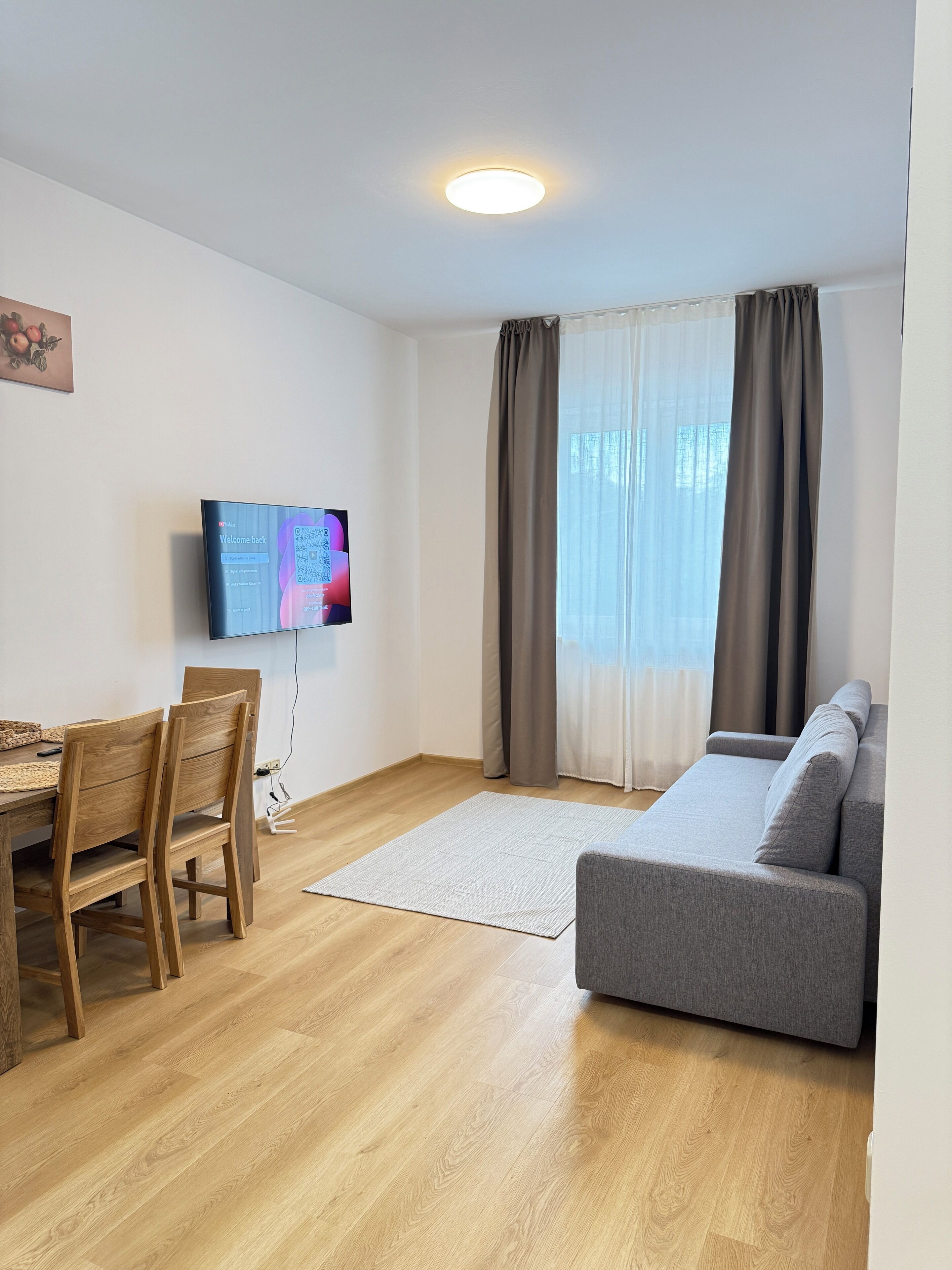 A living room with a grey sofa, wooden chairs, a flat-screen TV, a wooden dining table, and a window with curtains.